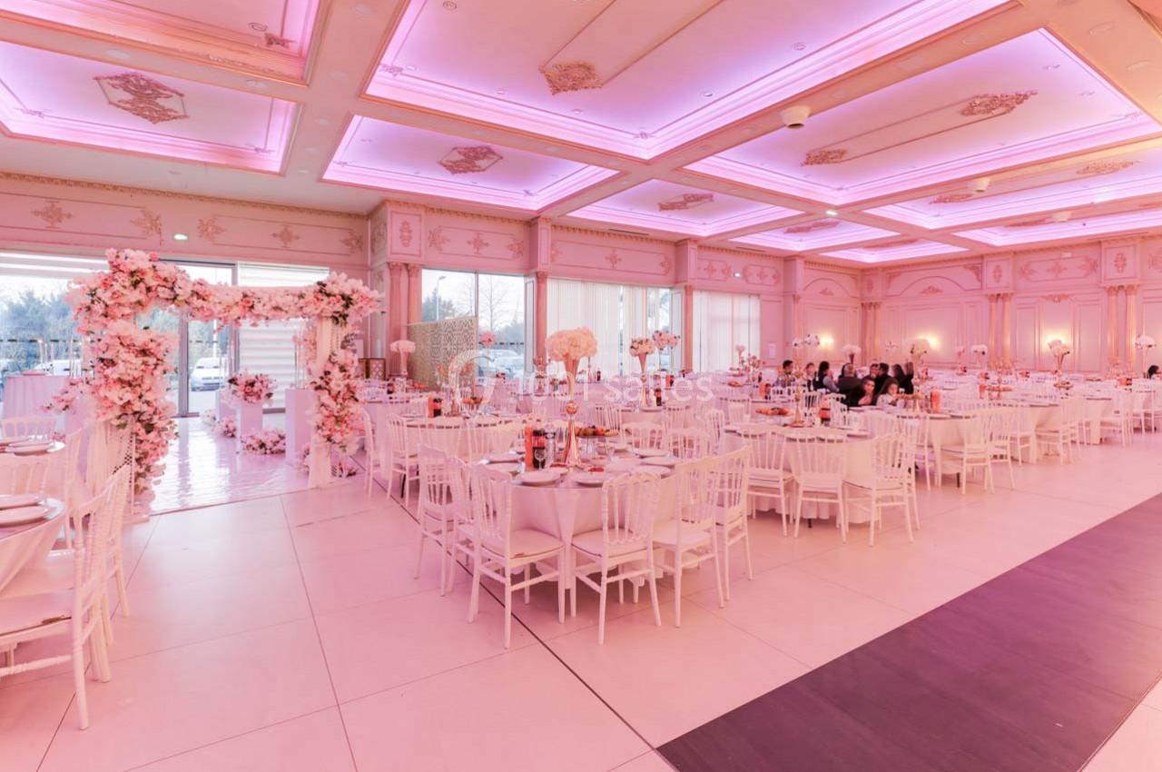 Salle de réception décorée avec des tons roses, tables dressées et arche florale près d'une entrée vitrée.