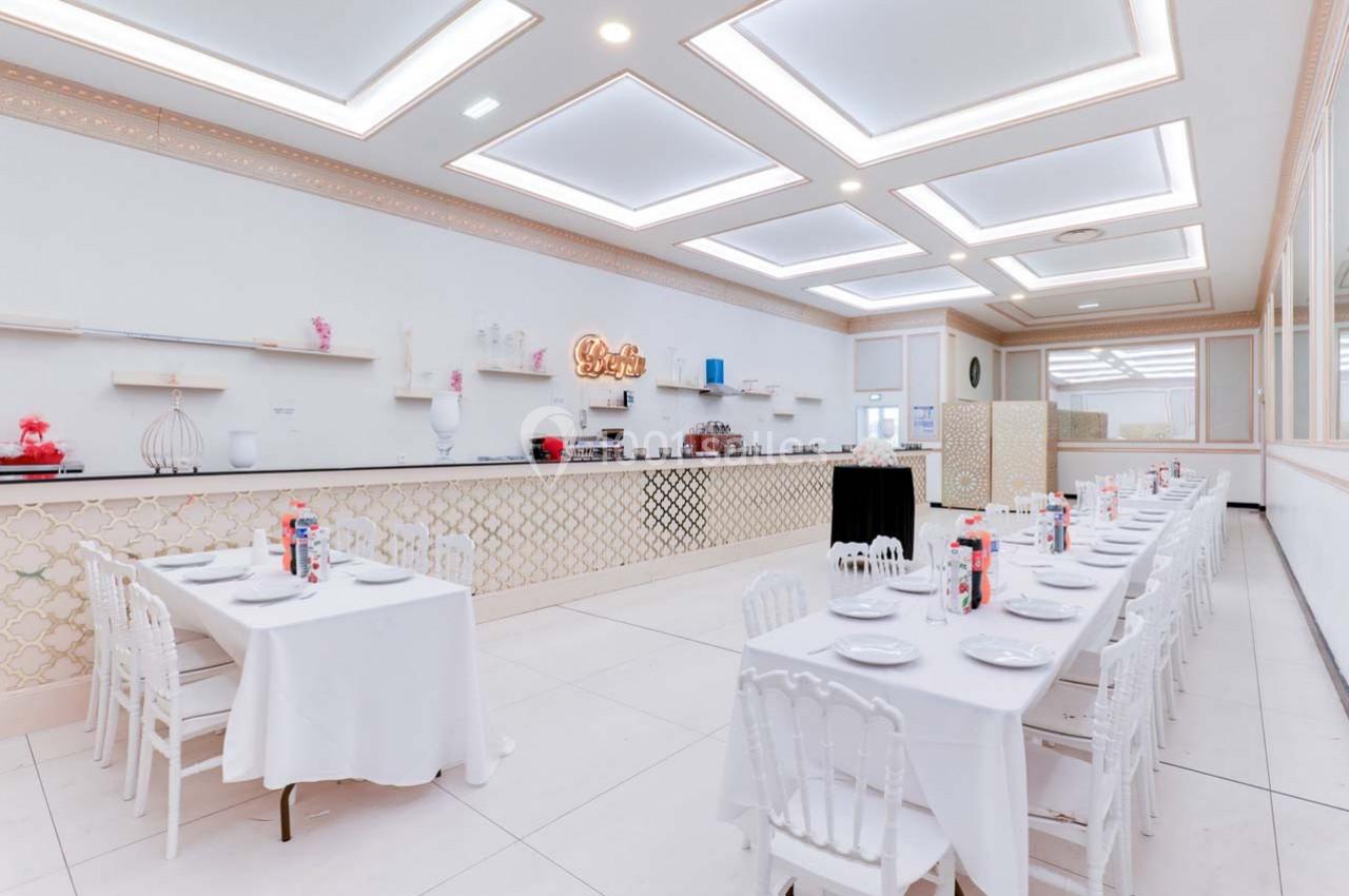 Salle lumineuse avec tables dressées, nappes blanches, chaises élégantes et comptoir décoratif en arrière-plan.