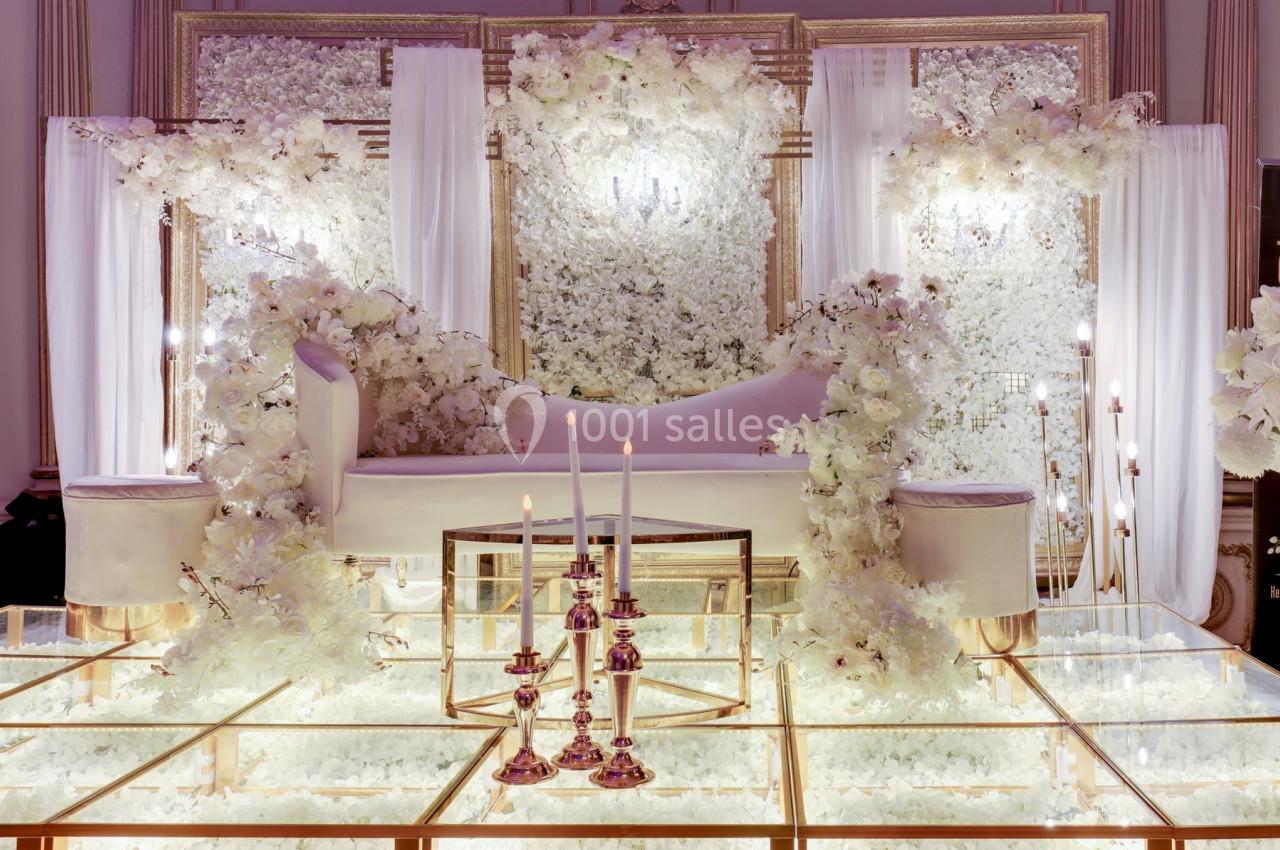 Décor élégant avec canapé blanc, fleurs blanches abondantes et chandeliers sur un sol en verre lumineux.