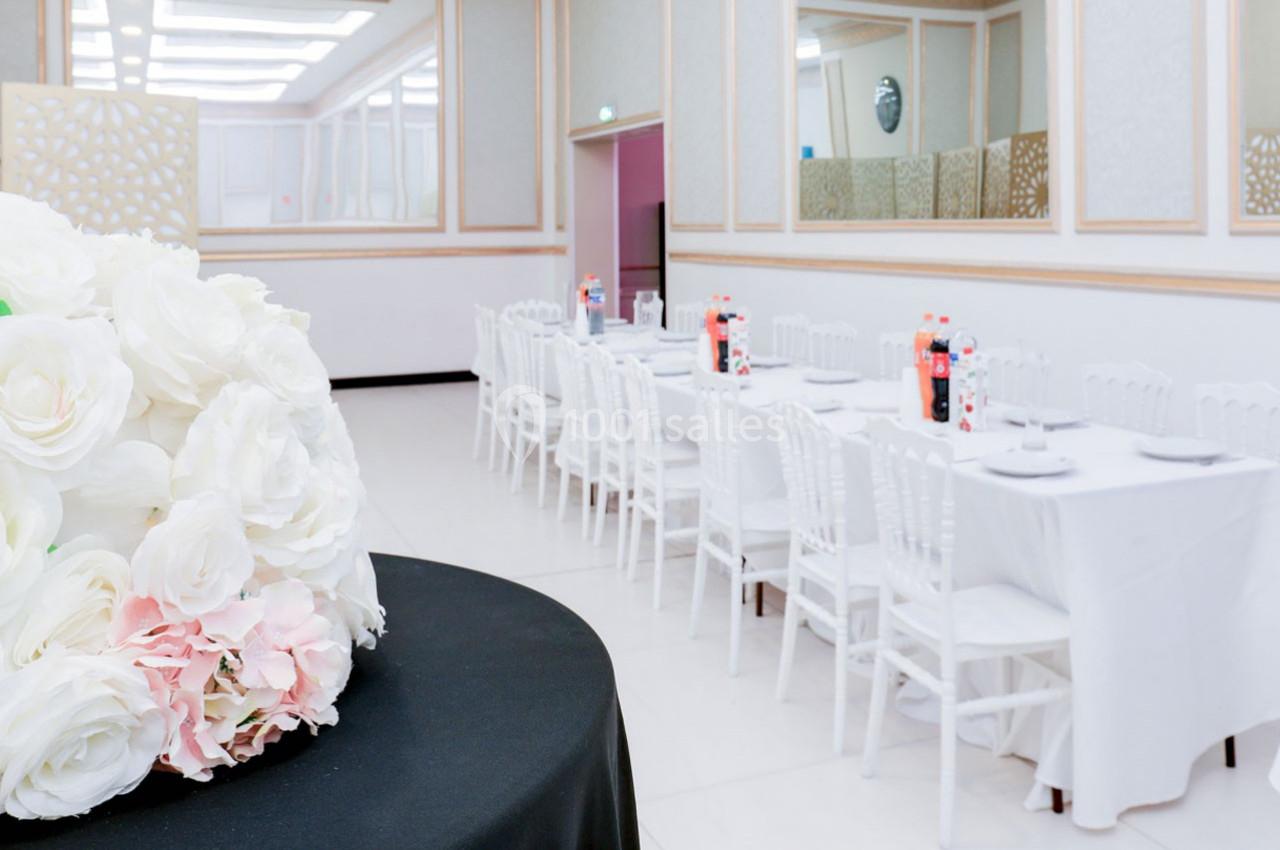 Salle de réception lumineuse avec tables dressées en blanc, décor floral au premier plan et bouteilles de soda visibles.