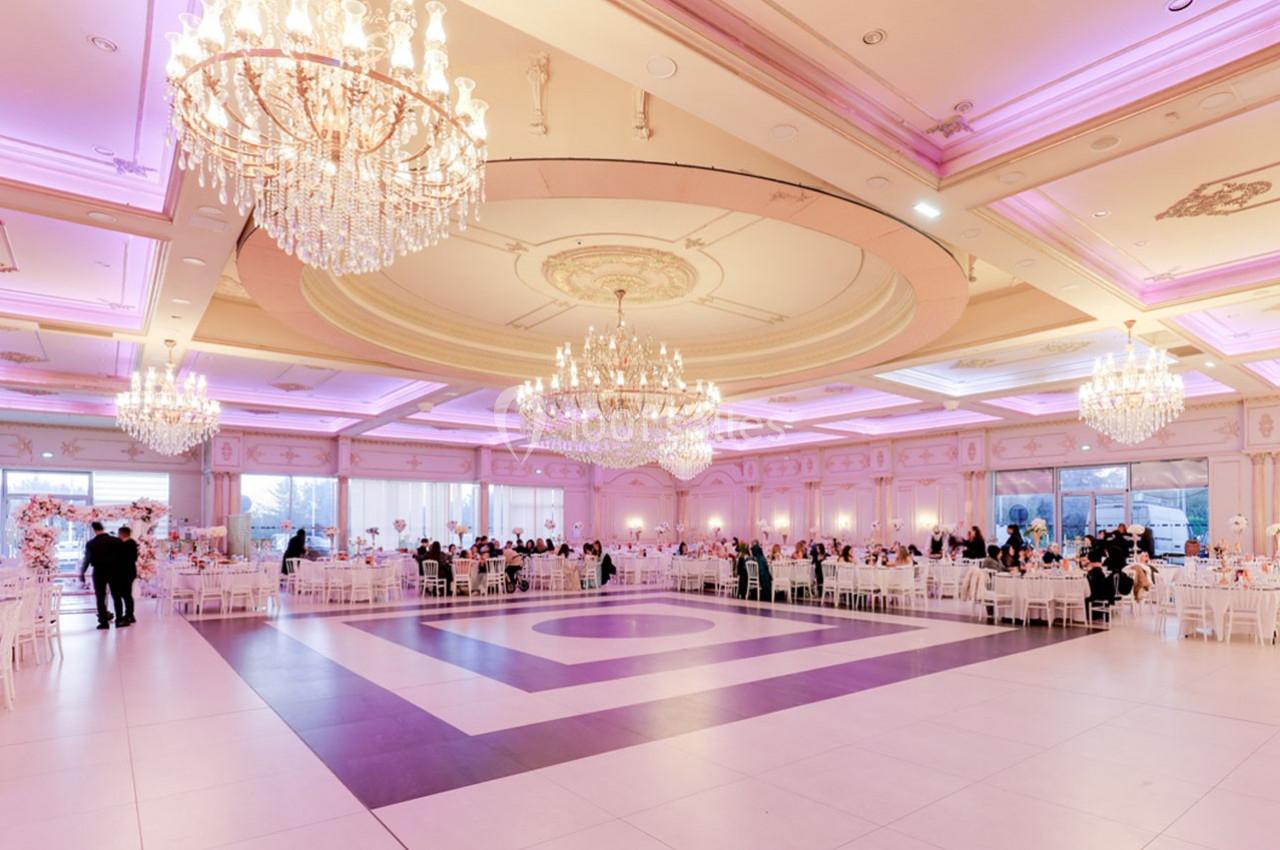 Salle de réception spacieuse avec des lustres imposants, des tables dressées et un éclairage rose tamisé.