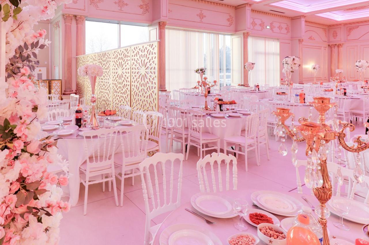 Salle de réception décorée en rose avec des tables rondes dressées, des chaises blanches et des ornements floraux.
