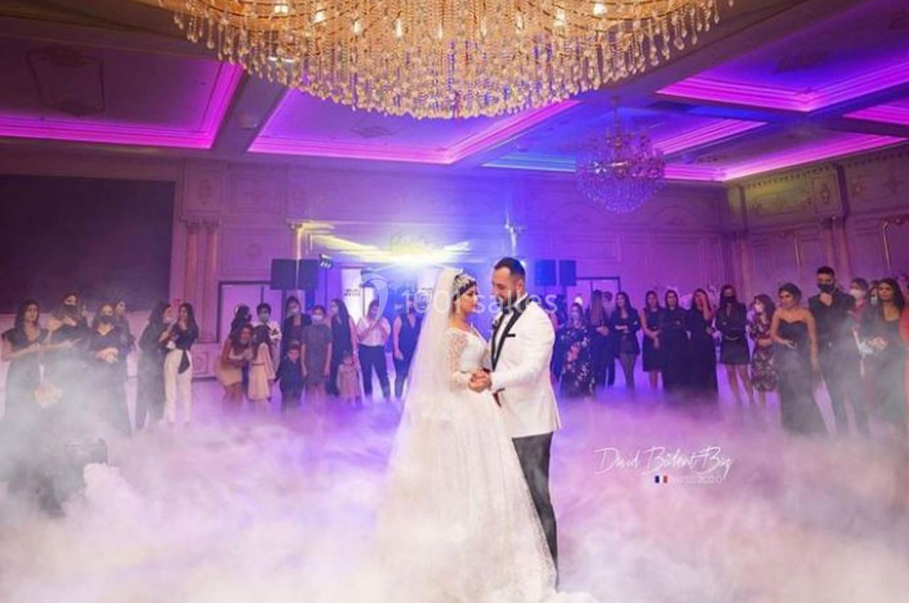 Un couple en tenue de mariage danse sous un lustre imposant, entouré de fumée et d'invités dans une salle éclairée.