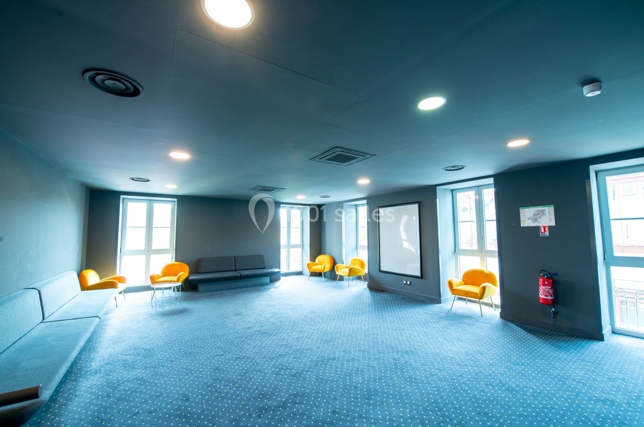 Salle lumineuse avec moquette bleue, fauteuils jaunes, canapés gris et écran mural, éclairée par des fenêtres.