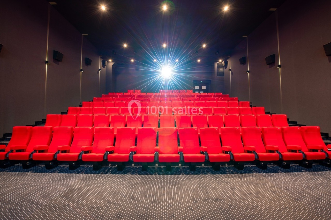 Salle de cinéma vide avec des rangées de fauteuils rouges et un projecteur allumé au fond.