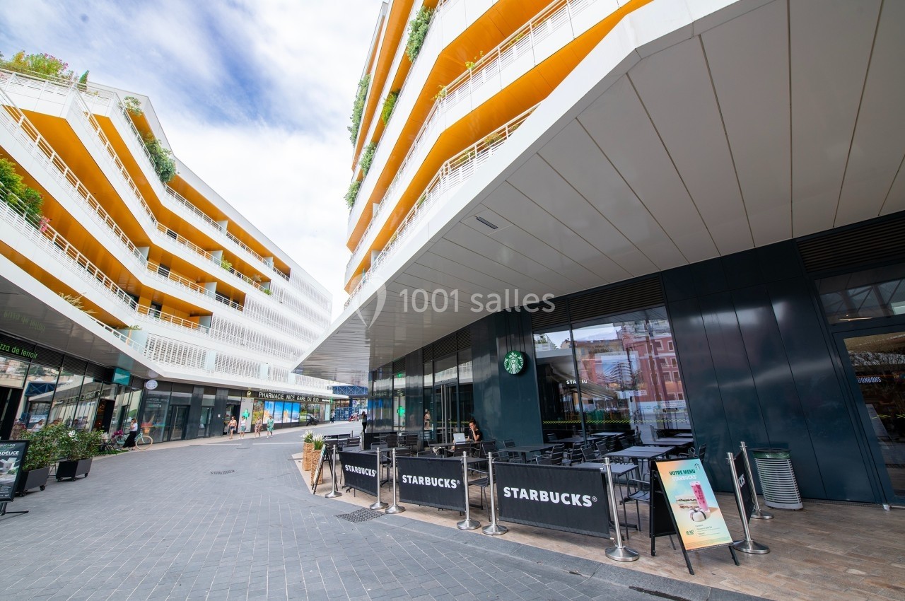 Terrasse d'un café Starbucks devant un immeuble moderne aux façades blanches et jaunes, sous un ciel partiellement nuageux.
