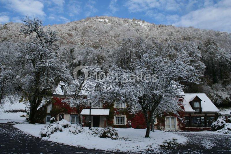Maison en pierre entourée d'arbres enneigés, au pied d'une colline boisée sous un ciel bleu.
