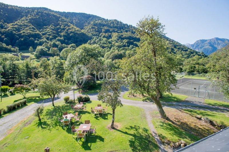 Vue d'un espace extérieur avec des tables sur une pelouse, entouré d'arbres et de montagnes en arrière-plan.