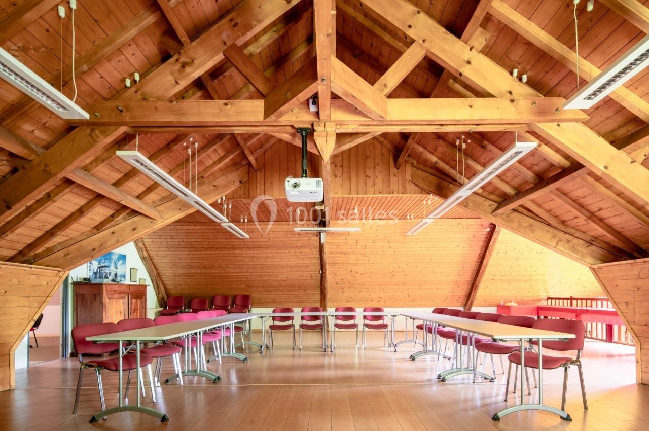 Salle de réunion lumineuse sous un toit en bois avec tables blanches, chaises rouges et projecteur suspendu.