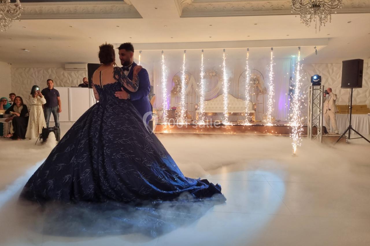 Un couple danse dans une salle de réception décorée, entouré de fumée au sol et de jets d'étincelles.