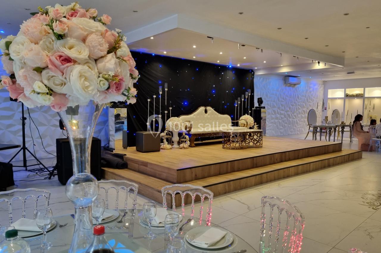 Salle de réception décorée avec un canapé sur une estrade, des chandeliers, des fleurs et des tables dressées.