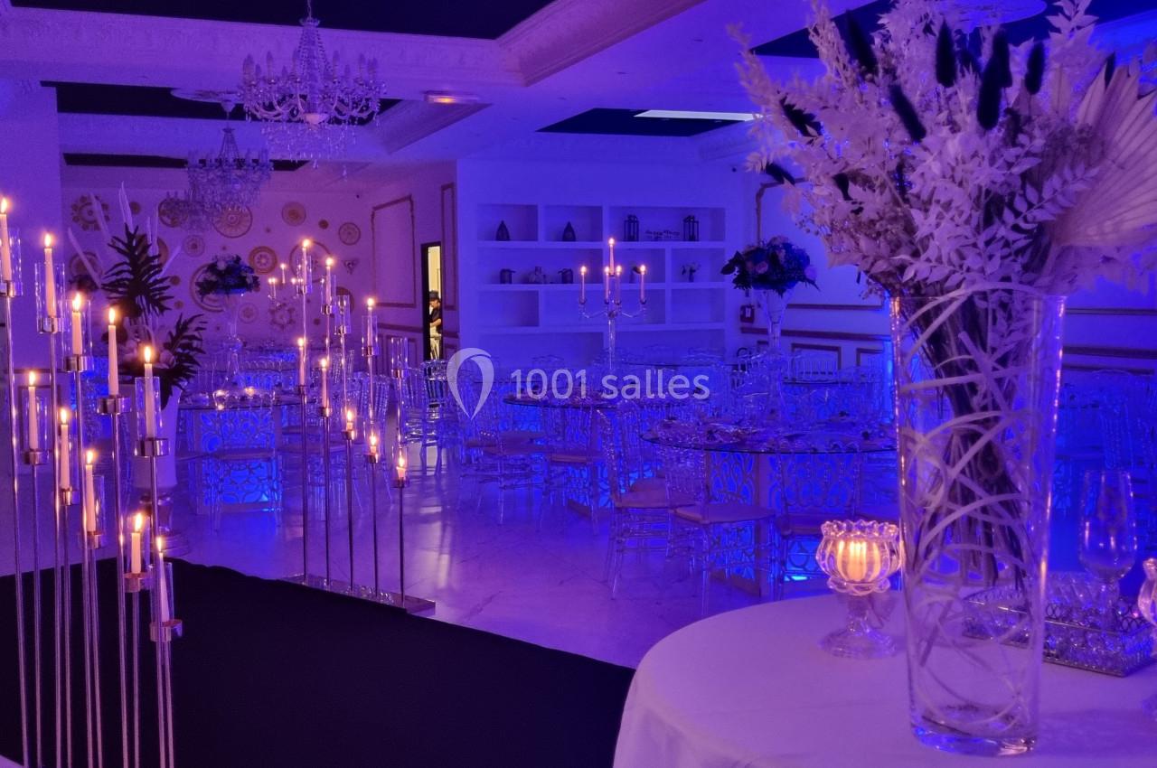 Salle de réception élégante avec éclairage bleu, chandeliers, tables décorées et arrangements floraux blancs.