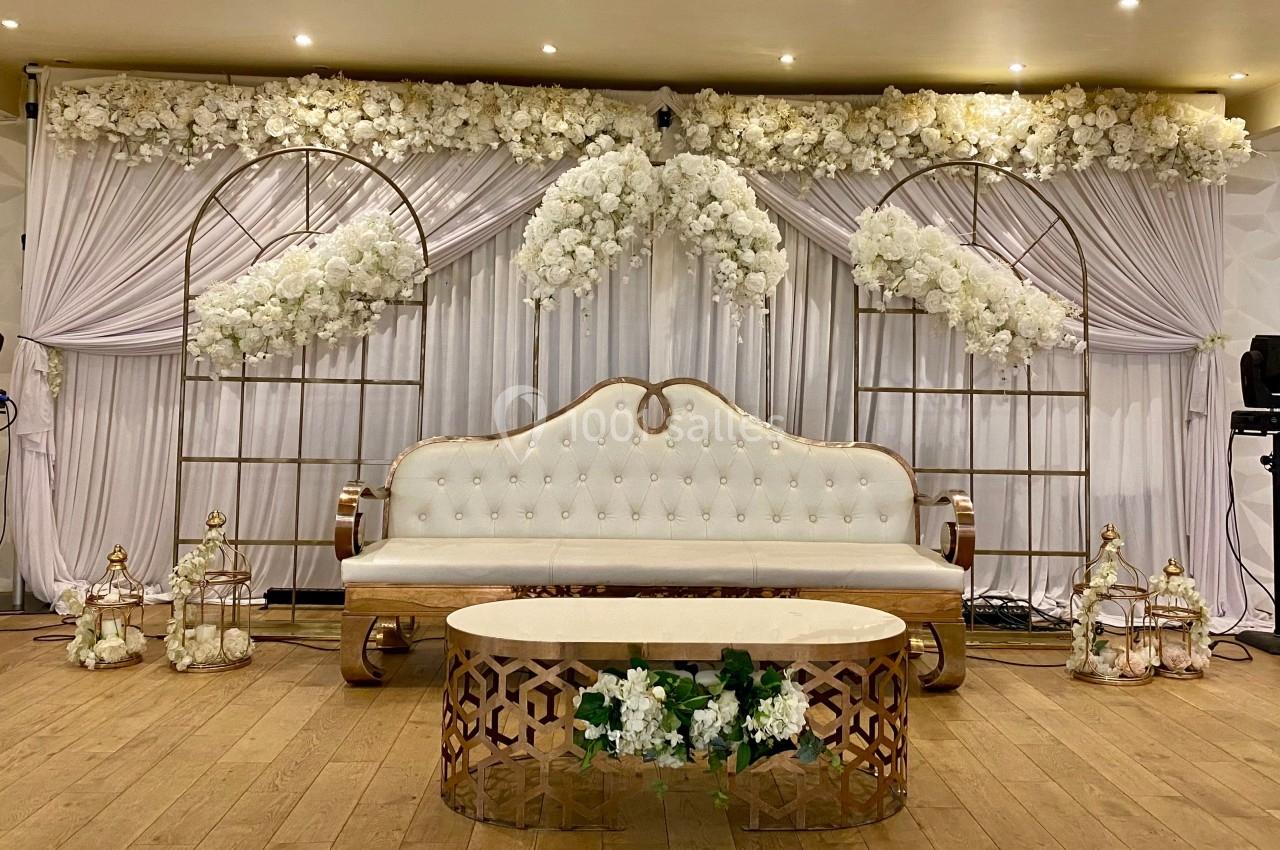 Décor de mariage avec canapé blanc capitonné, rideaux et arches ornés de fleurs blanches, table basse en bois.