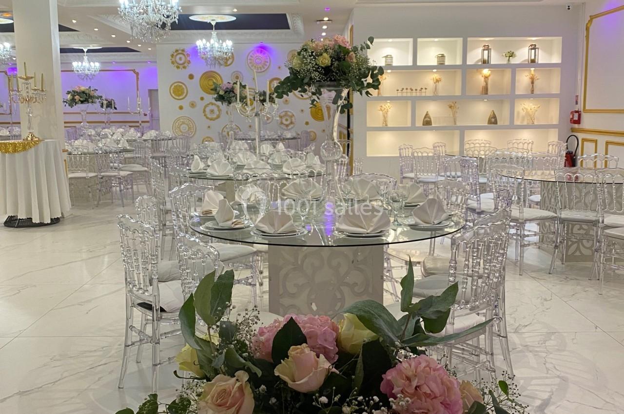 Salle de réception élégante avec tables dressées, chaises transparentes et décorations florales dans un cadre lumineux.