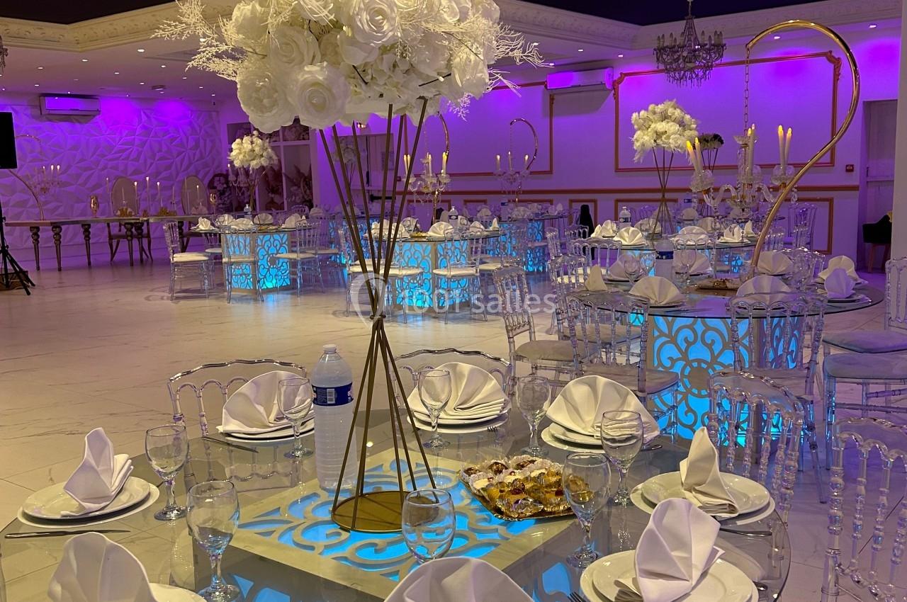 Salle de réception élégante avec tables rondes en verre, décor floral blanc et éclairage violet.