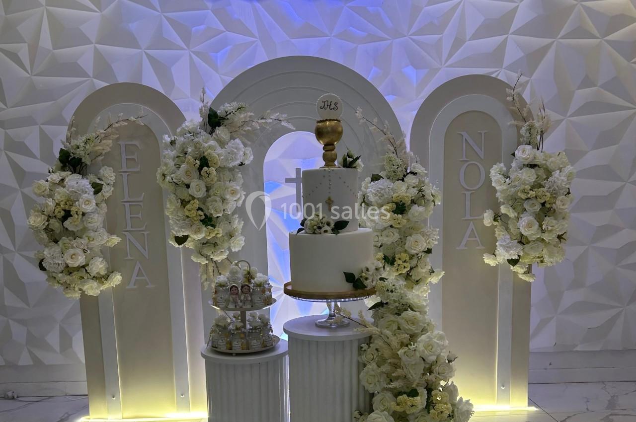 Décoration de cérémonie avec fleurs blanches, arche lumineuse, gâteau à étages et calice doré au centre.