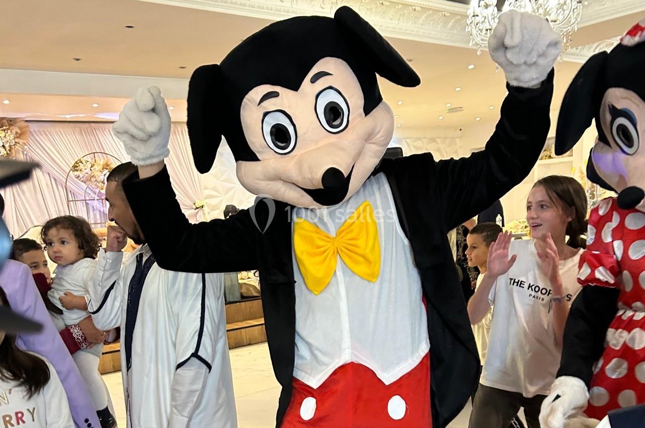 Personne déguisée en souris anthropomorphe avec un nœud jaune et un costume rouge, entourée d'enfants dans un lieu festif.