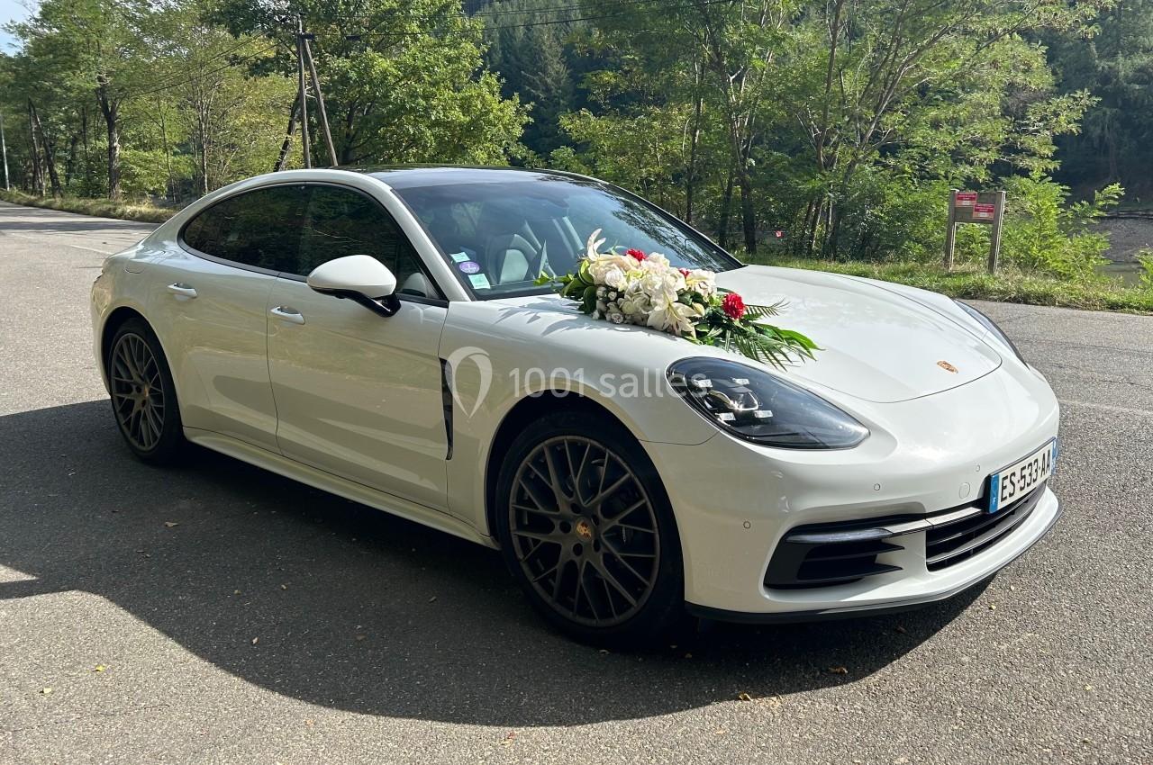 Voiture blanche décorée de fleurs blanches et vertes, stationnée sur une route bordée d'arbres.