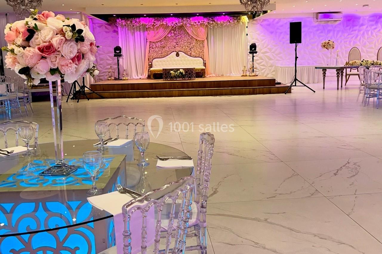 Salle de réception élégante avec décorations florales, chaises transparentes et scène ornée de rideaux et lumières.