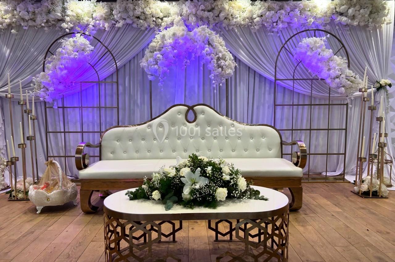 Canapé blanc capitonné devant des rideaux et des arches décorés de fleurs blanches, table basse ornée d'un bouquet.