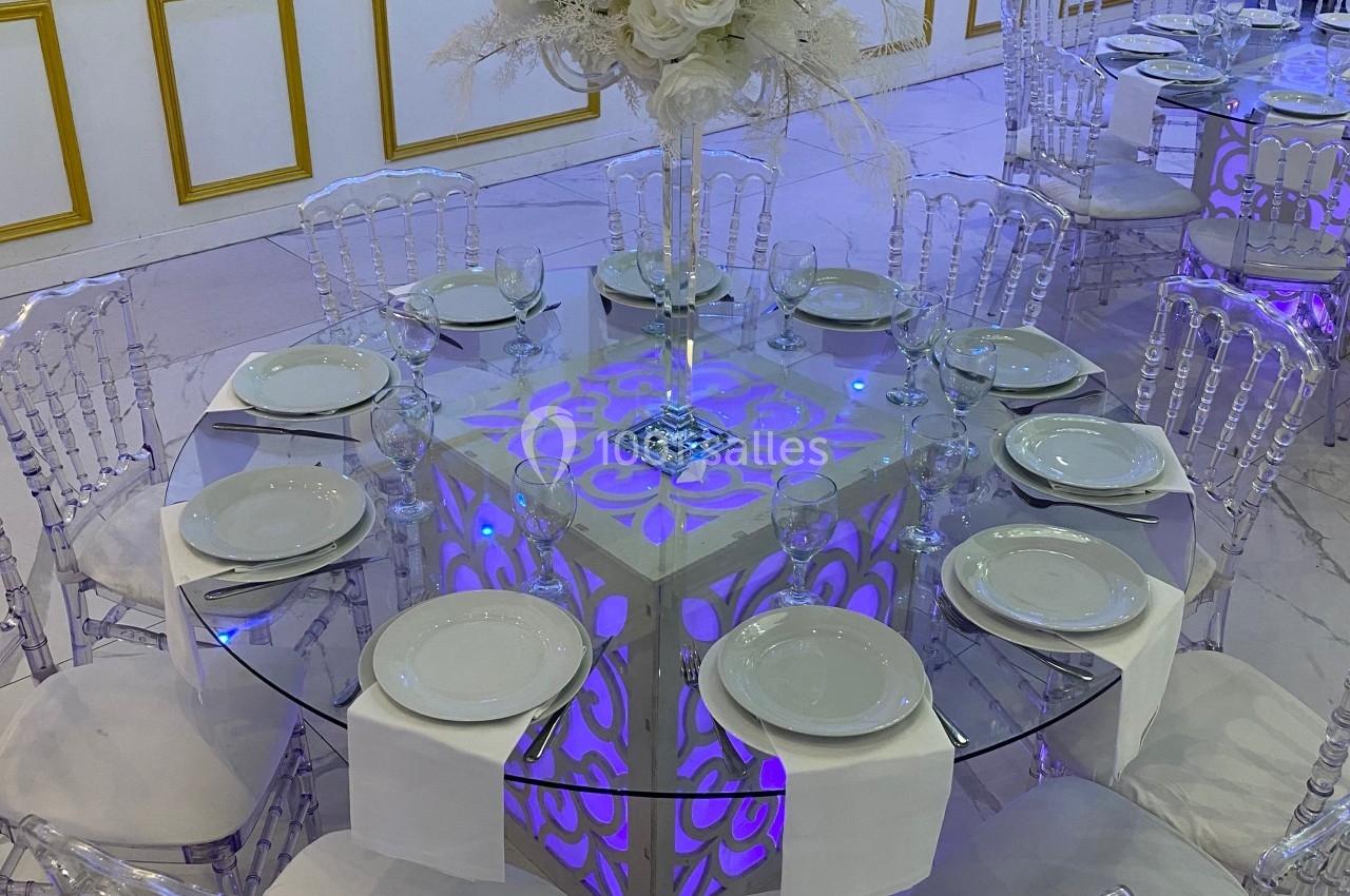 Table décorée pour un événement avec nappes blanches, assiettes, chaises transparentes et centre de table floral blanc.
