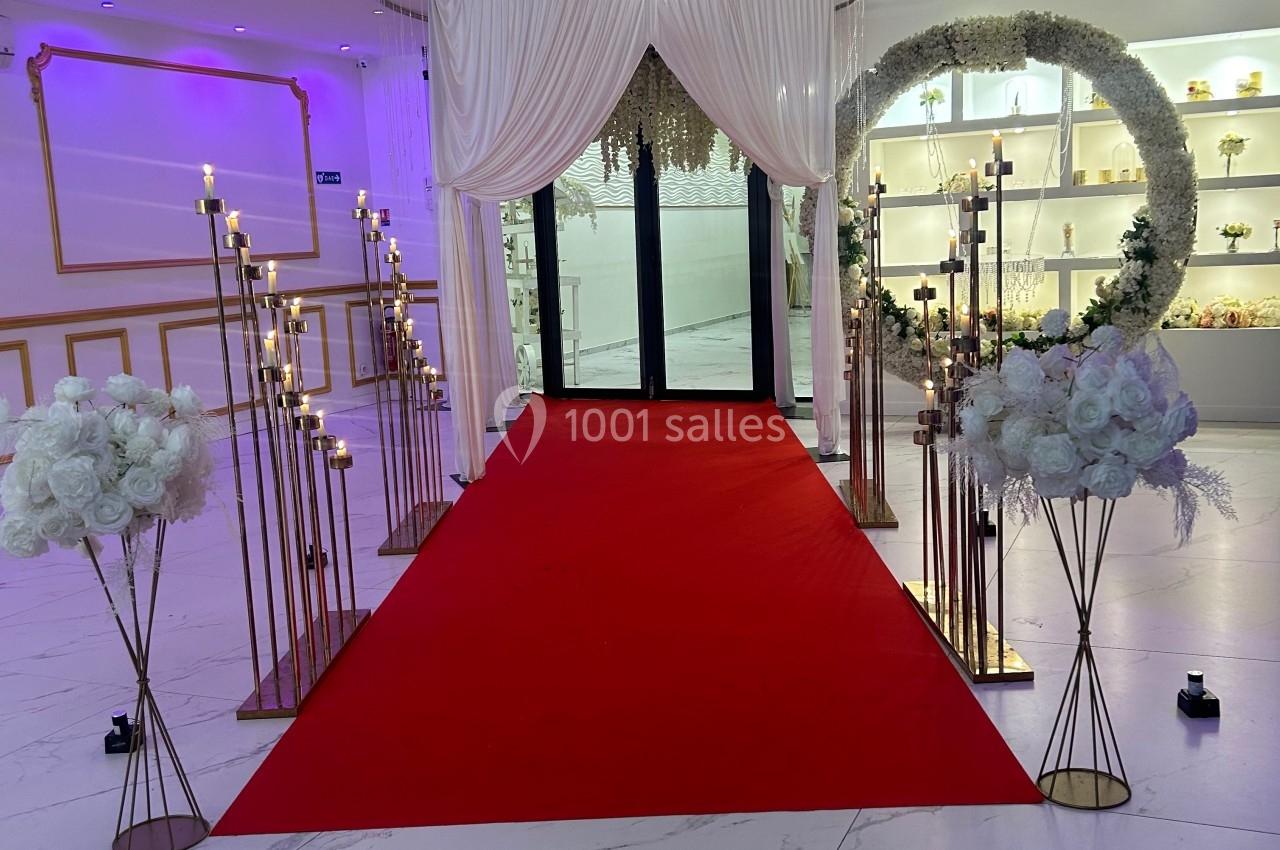 Allée décorée de chandeliers et de fleurs blanches menant à une porte, avec un tapis rouge et des rideaux blancs.