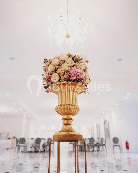 Vase doré rempli de fleurs roses et blanches, posé sur un support, dans une salle lumineuse avec lustre et tables en arrière…