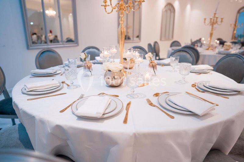 Table ronde élégamment dressée avec nappes blanches, couverts dorés, verres en cristal et centre de table floral.