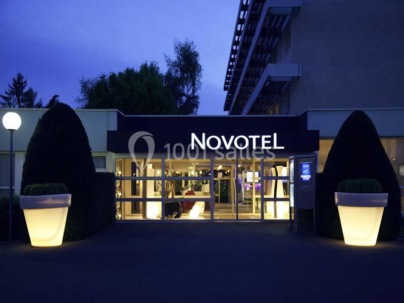 Entrée principale d'un hôtel Novotel éclairée en soirée, avec des arbustes taillés et de grands pots lumineux.