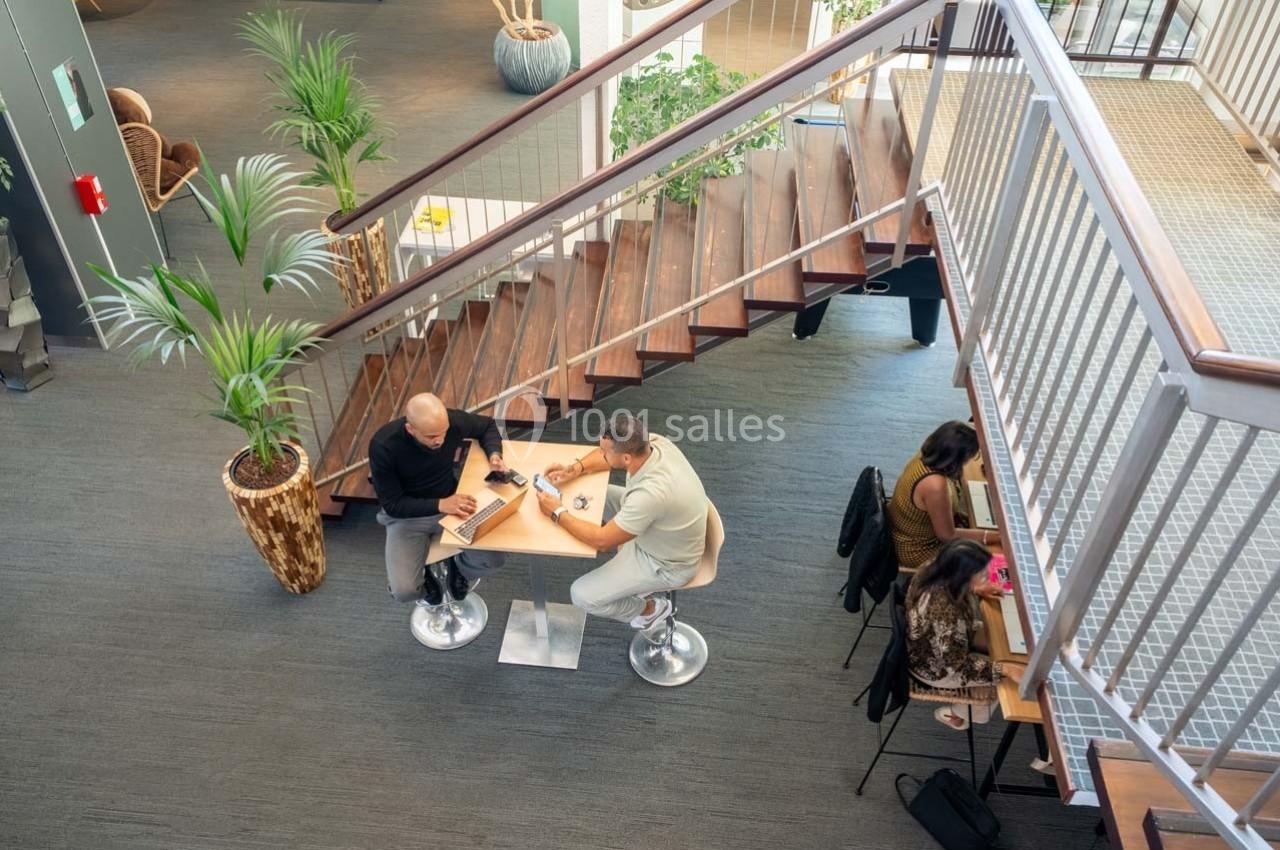 Deux hommes discutent à une table haute dans un espace de travail moderne avec escalier et plantes.