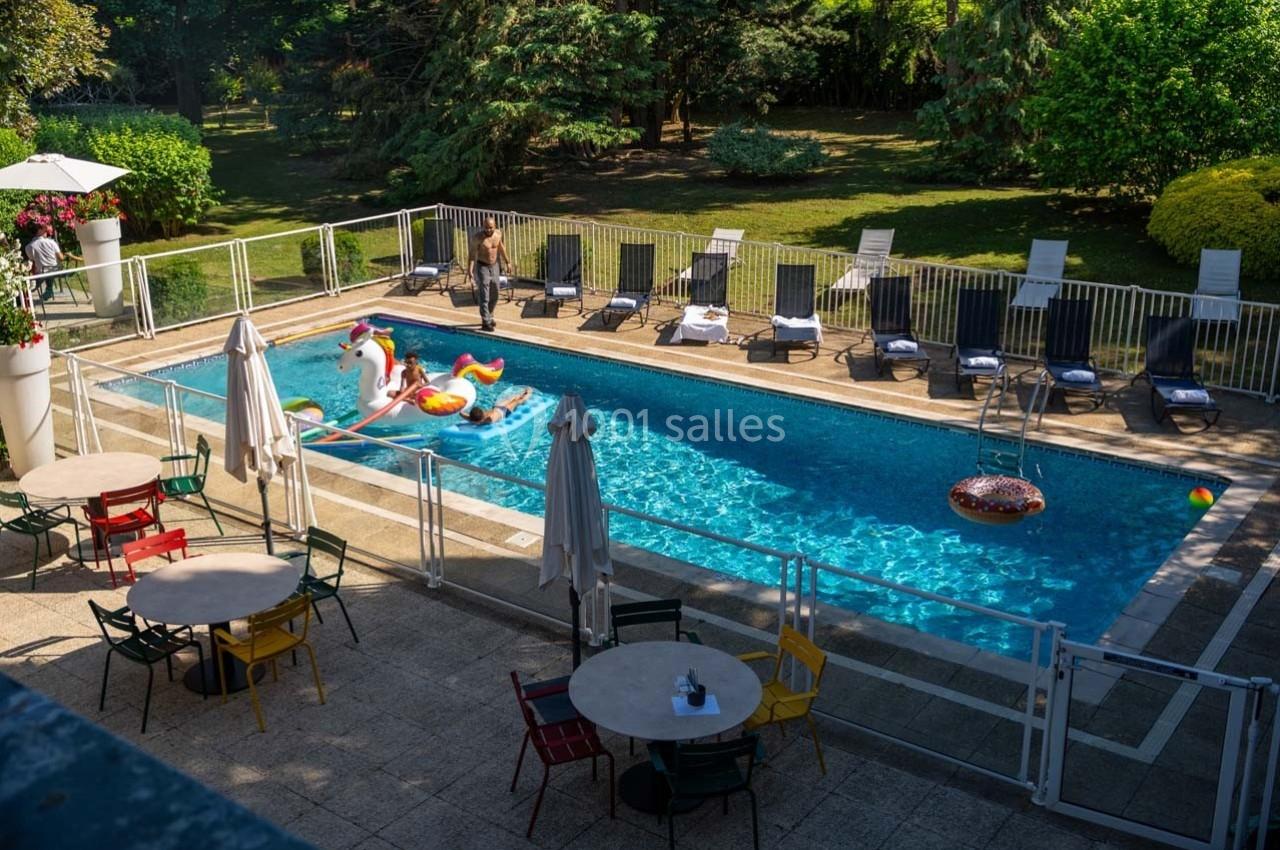 Piscine extérieure entourée de chaises longues, avec des bouées colorées flottant sur l'eau et un jardin en arrière-plan.