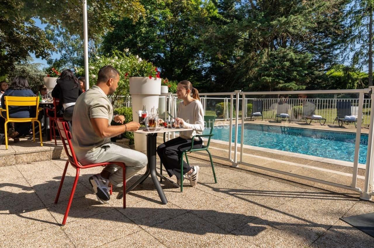 Deux personnes dînent en terrasse près d'une piscine, entourées de chaises colorées et de végétation.