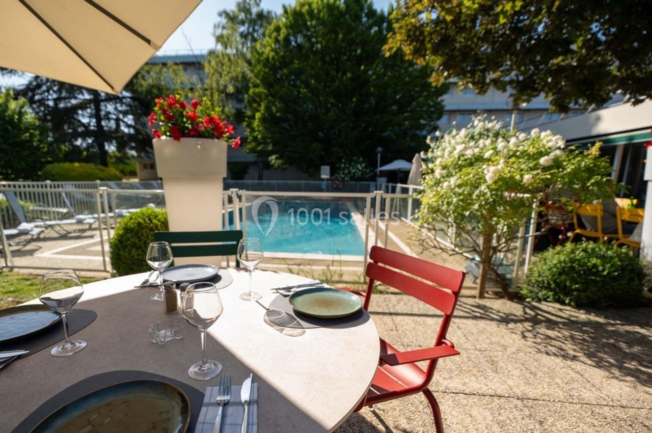 Table dressée en terrasse avec vue sur une piscine entourée de végétation et de chaises colorées.