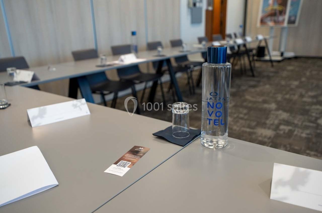 Salle de réunion avec tables disposées en U, bouteilles d'eau, verres, papiers et supports pour participants.