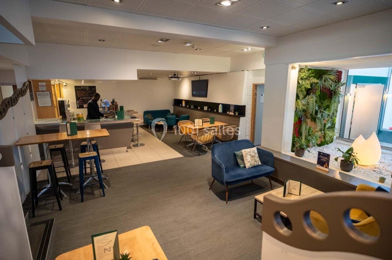 Espace lounge lumineux avec tables, chaises hautes, fauteuils, mur végétal et comptoir de service en arrière-plan.