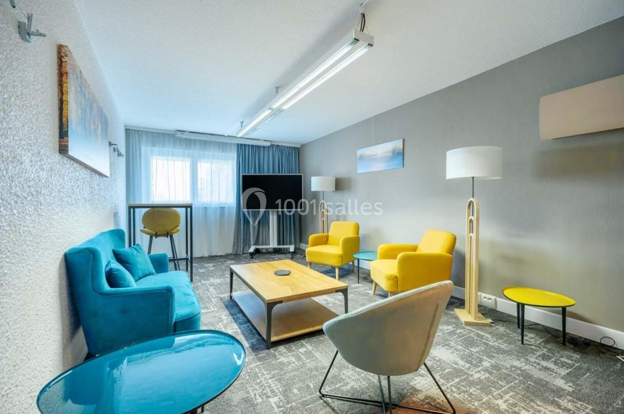 Salle de réunion lumineuse avec canapés bleus, fauteuils jaunes, table basse en bois et écran de télévision.