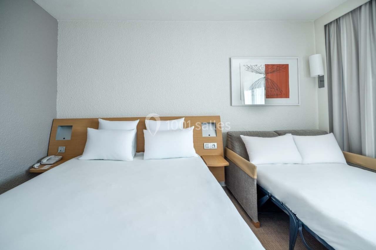 Chambre d'hôtel avec un lit double, un canapé-lit, des murs clairs et un tableau encadré au mur.