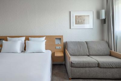 Chambre avec un lit double, un canapé gris, un mur clair et un tableau encadré.