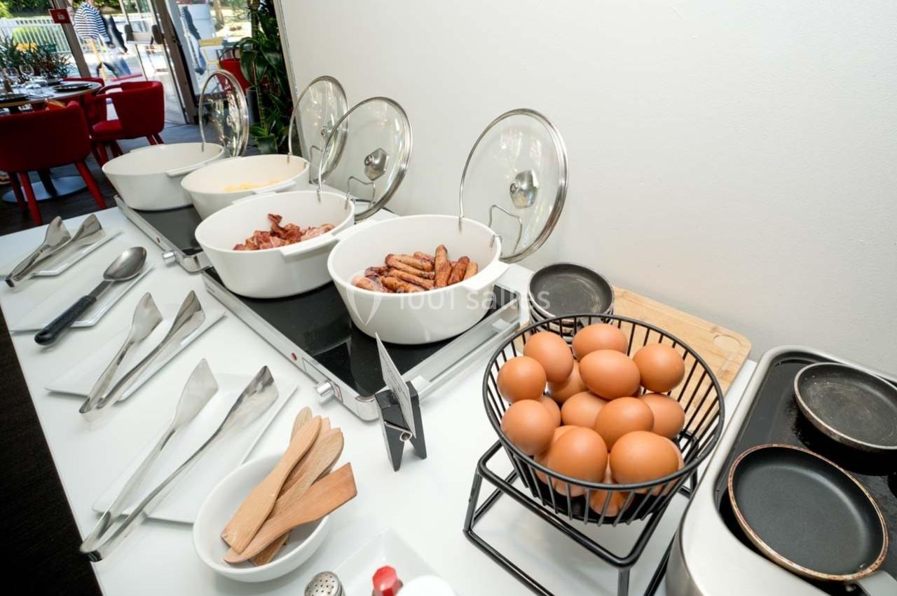 Buffet de petit-déjeuner avec œufs, saucisses, bacon et ustensiles disposés sur une table blanche.