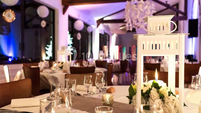Salle de réception décorée avec des tables dressées, des lanternes blanches et un éclairage tamisé.