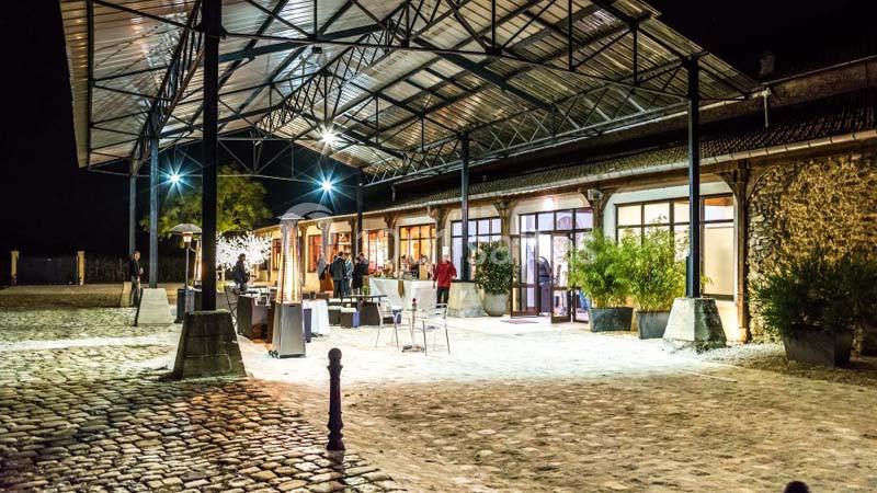 Vue nocturne d'un bâtiment avec une structure métallique, éclairé, entouré de pavés et de plantes en pot.