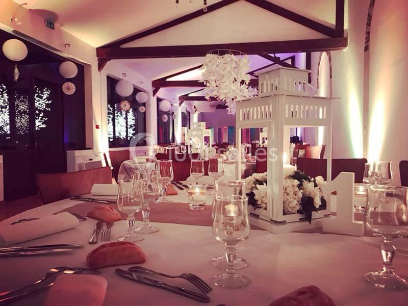 Salle de réception décorée avec des tables dressées, lanternes blanches, fleurs et éclairage tamisé.