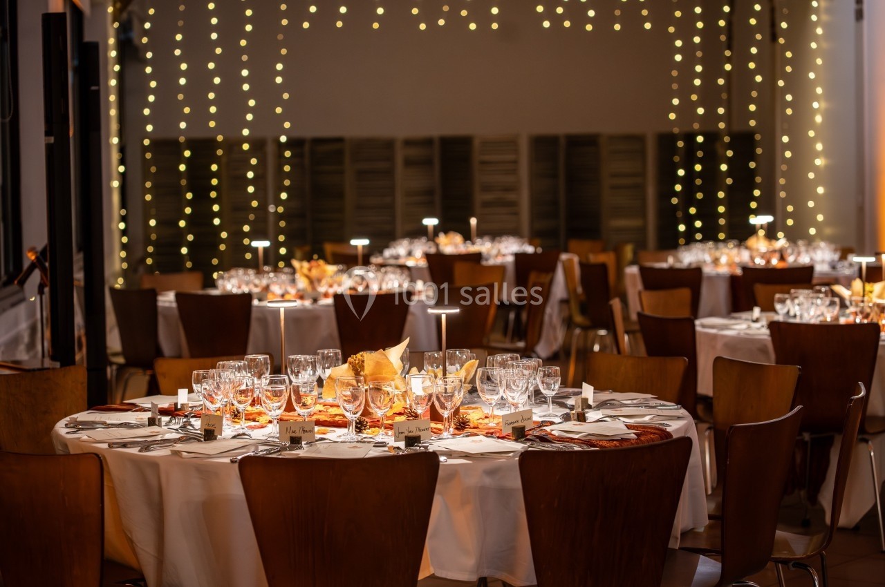 Salle de réception décorée avec des tables rondes, nappes blanches, vaisselle élégante et guirlandes lumineuses suspendues.