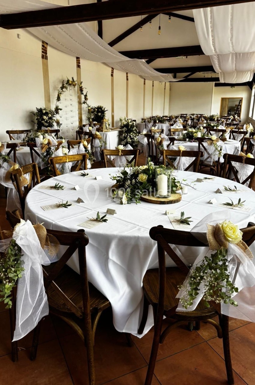 Salle de réception décorée pour un mariage, avec tables rondes ornées de nappes blanches, fleurs et bougies.