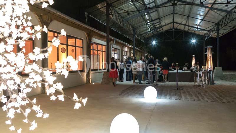 Un groupe de personnes discute près d'un bar sous un auvent éclairé, avec des décorations lumineuses au premier plan.