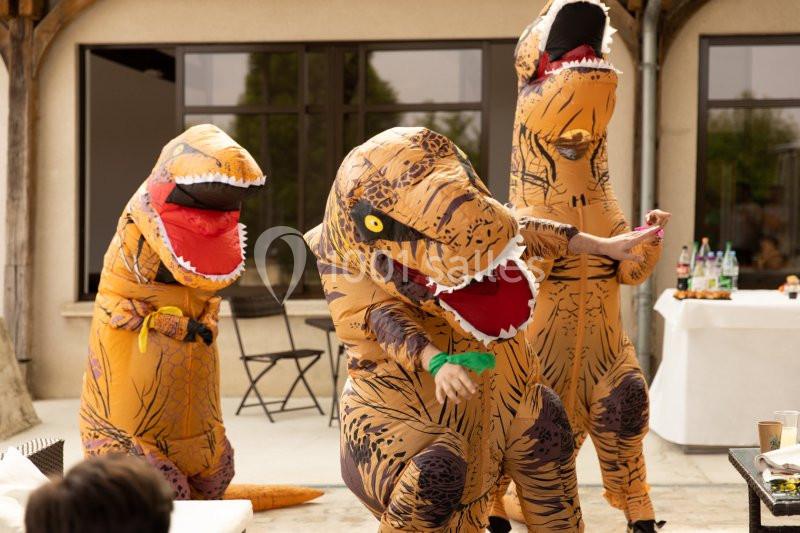 Trois personnes déguisées en dinosaures dansent dans une cour extérieure près de tables et chaises.