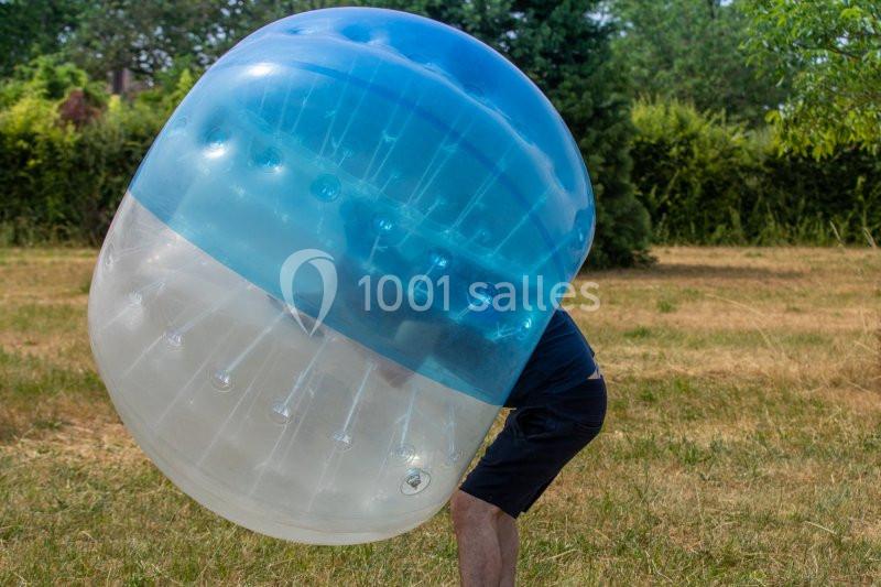 Personne portant une bulle gonflable transparente et bleue dans un espace extérieur herbeux.