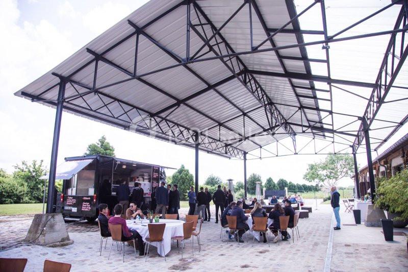 Un groupe de personnes assises et debout sous un grand auvent près d'un food truck dans un espace extérieur.