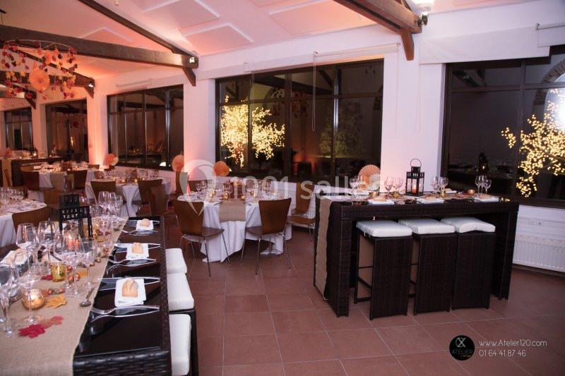 Salle de réception décorée pour un événement, avec tables dressées, chaises en bois et éclairage chaleureux.