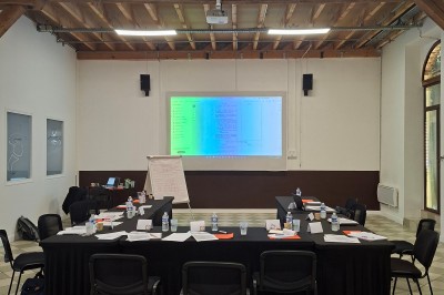 Salle de réunion avec une grande table rectangulaire, chaises noires, documents, bouteilles d'eau et écran de présentation.